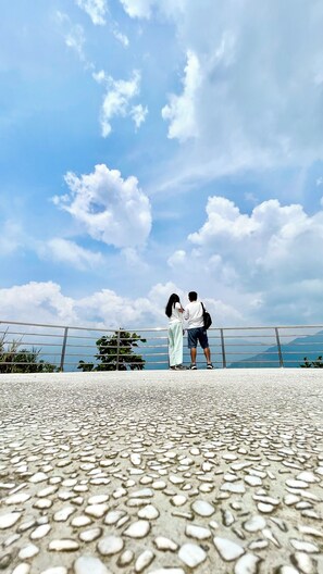 A couple standing on a paved area with a metal railing, overlooking a body of water and distant mountains under a partly cloudy sky.