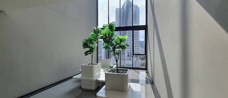 A modern interior with a high ceiling, a glass wall with a cityscape view, and two potted plants on a reflective floor.