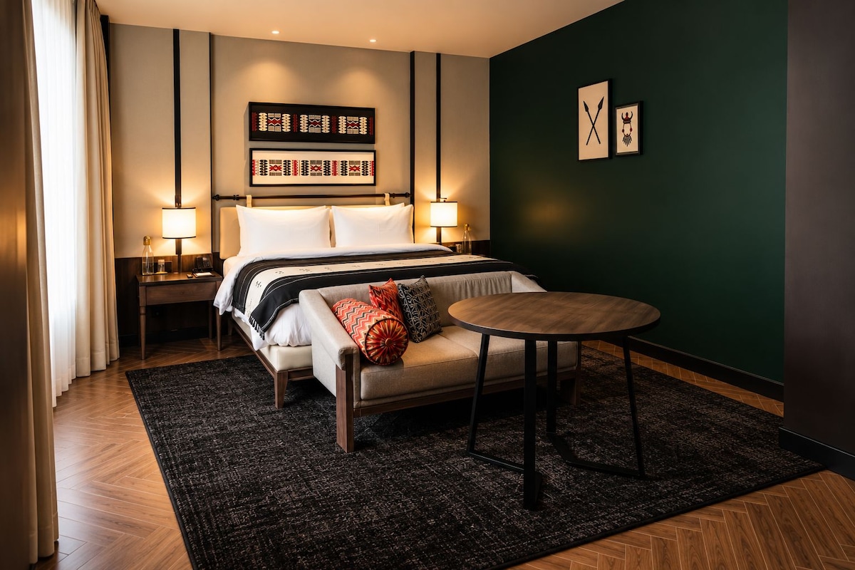 A hotel room with a bed, bedside tables, a round table, and a dark green accent wall.