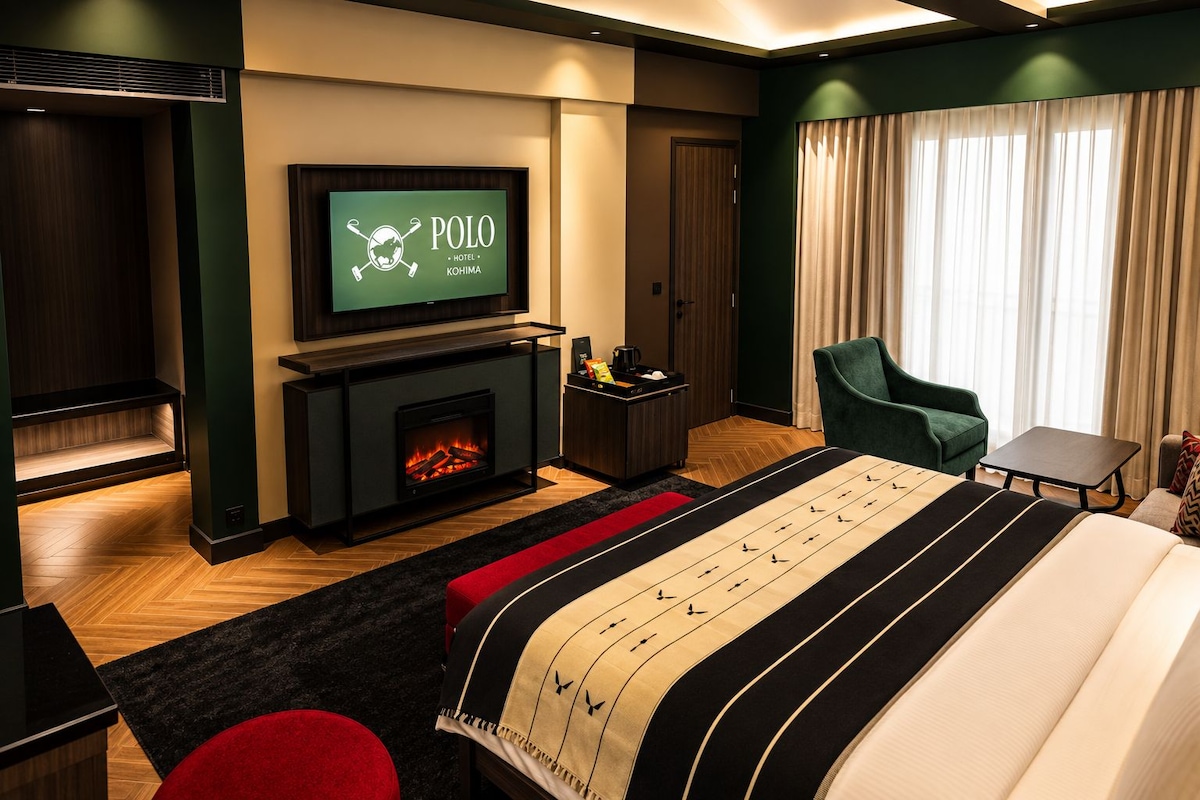 A hotel room with a television displaying the Polo Hotel logo, a fireplace, a bed with a striped blanket, a green armchair, and a small table.