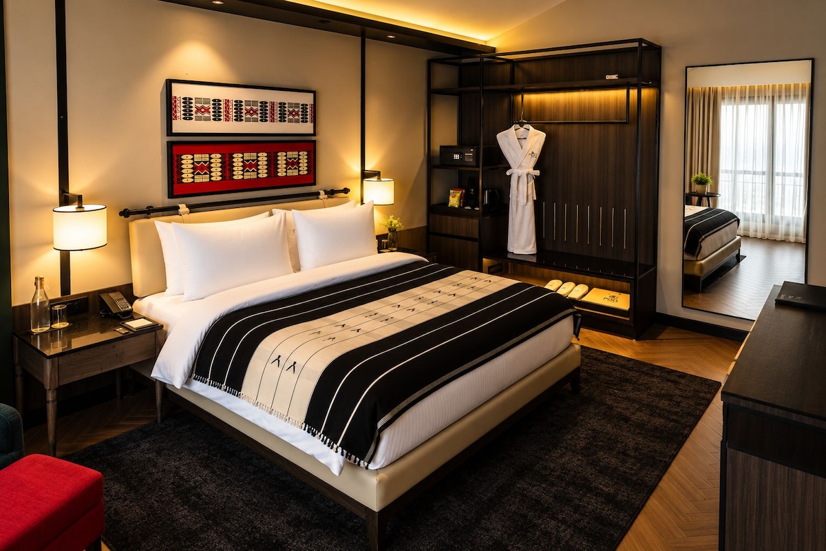 A hotel room with a large bed, bedside lamps, a robe hanging on a rack, a mirror, and a view of the room through a window.