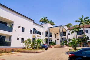 Un edificio de varios pisos con entrada cubierta, palmeras y coches aparcados.