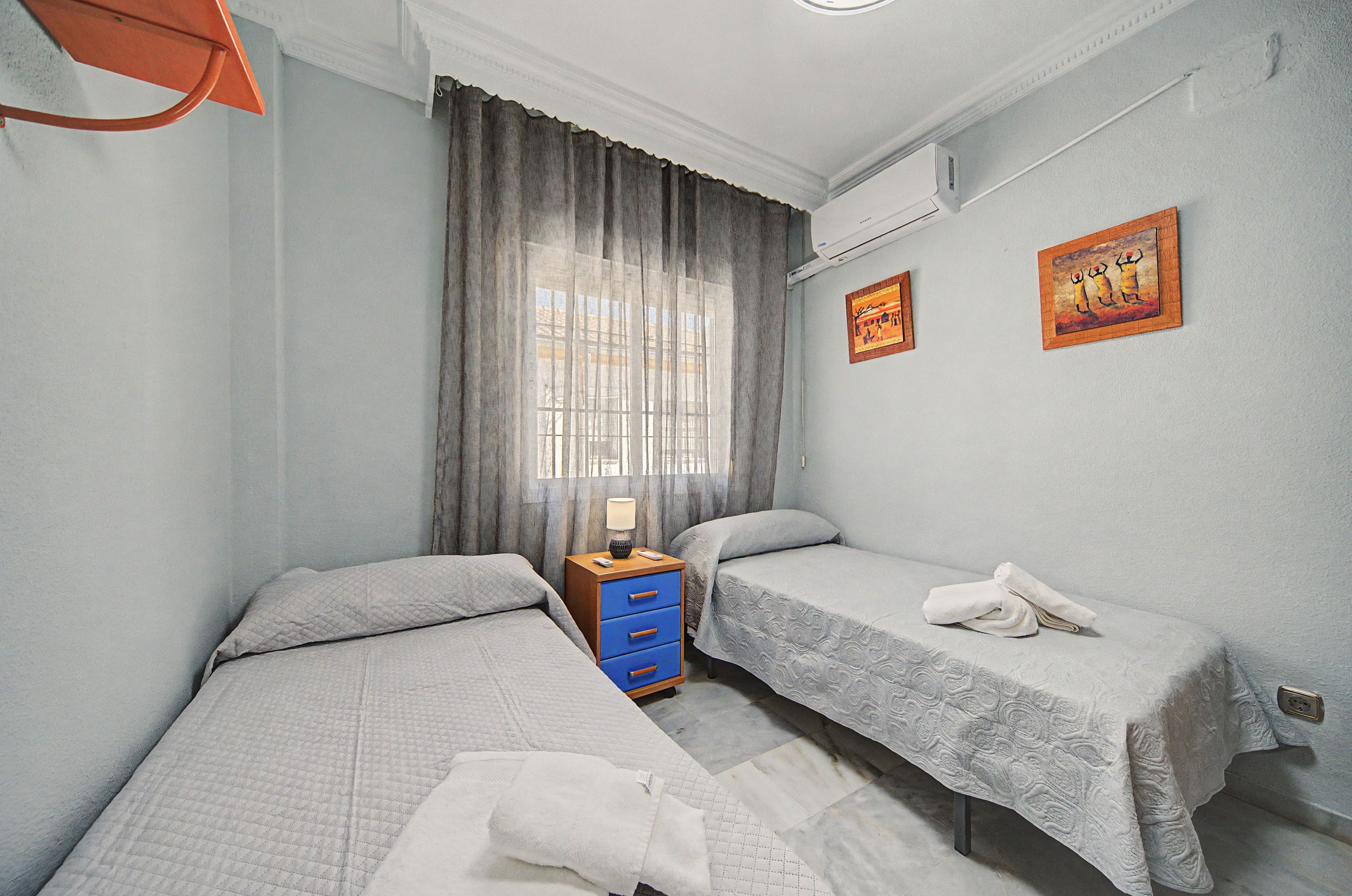 Hotellihuone, jossa on kaksi erillistä sänkyä, yöpöytä, lamppu ja kaksi seinälle ripustettua taulua.