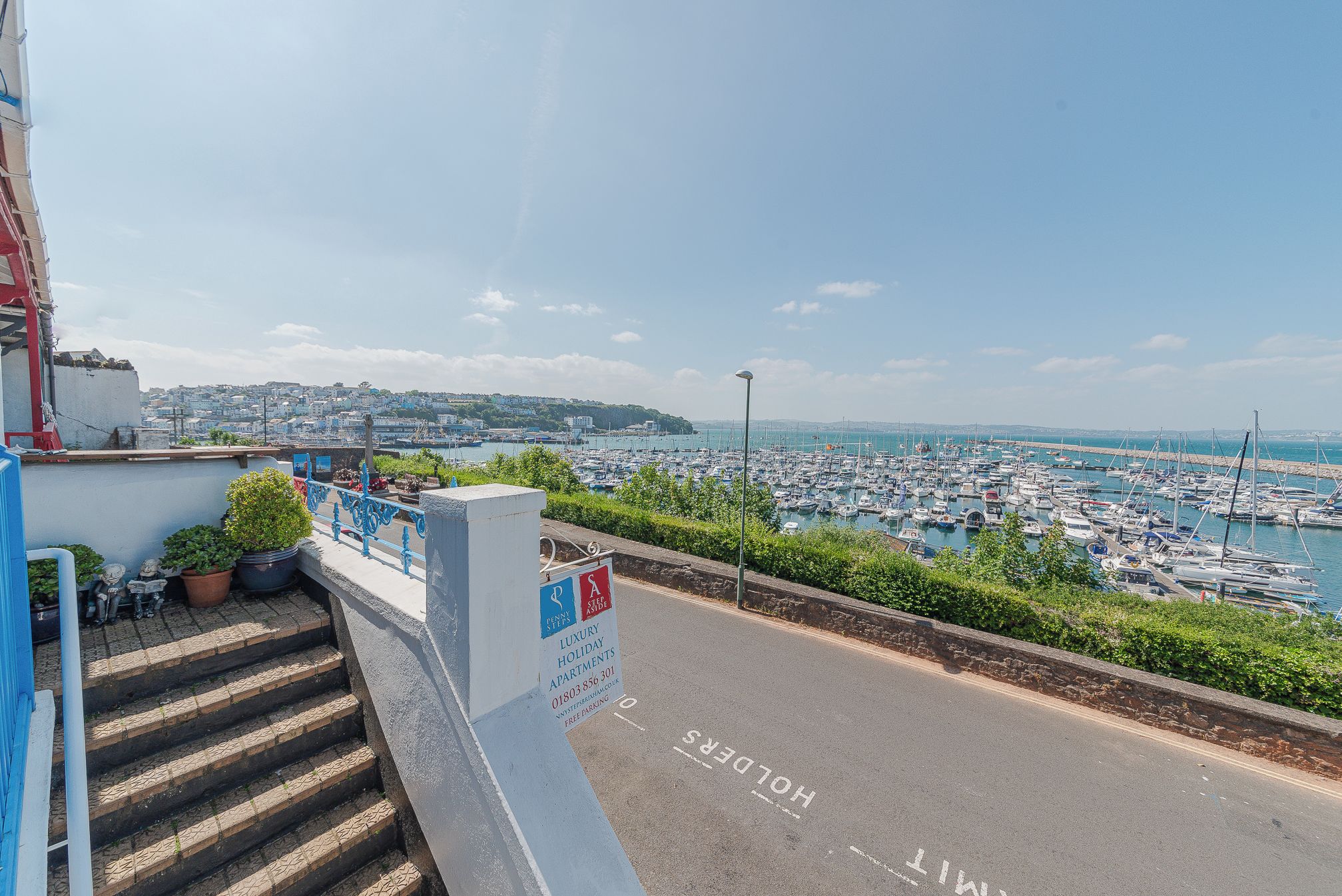 A staircase leading to a balcony overlooking a marina with boats, a sign for luxury holiday apartments, and a clear view of the town in the distance.