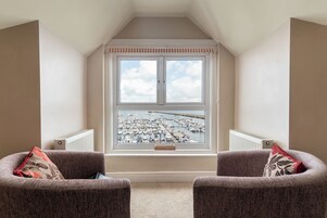 Una habitación con ventanal que da a un puerto deportivo, dos sillones con cojines y un radiador.