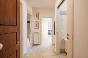 Un couloir avec une porte en bois, un radiateur et un sol carrelé, qui mène à une pièce dotée d’une fenêtre.