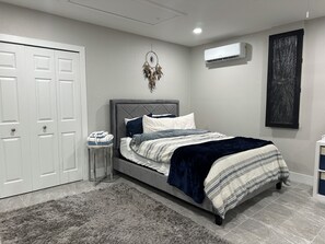 A bedroom with a bed, a bedside table, a wall-mounted air conditioner, and a door.