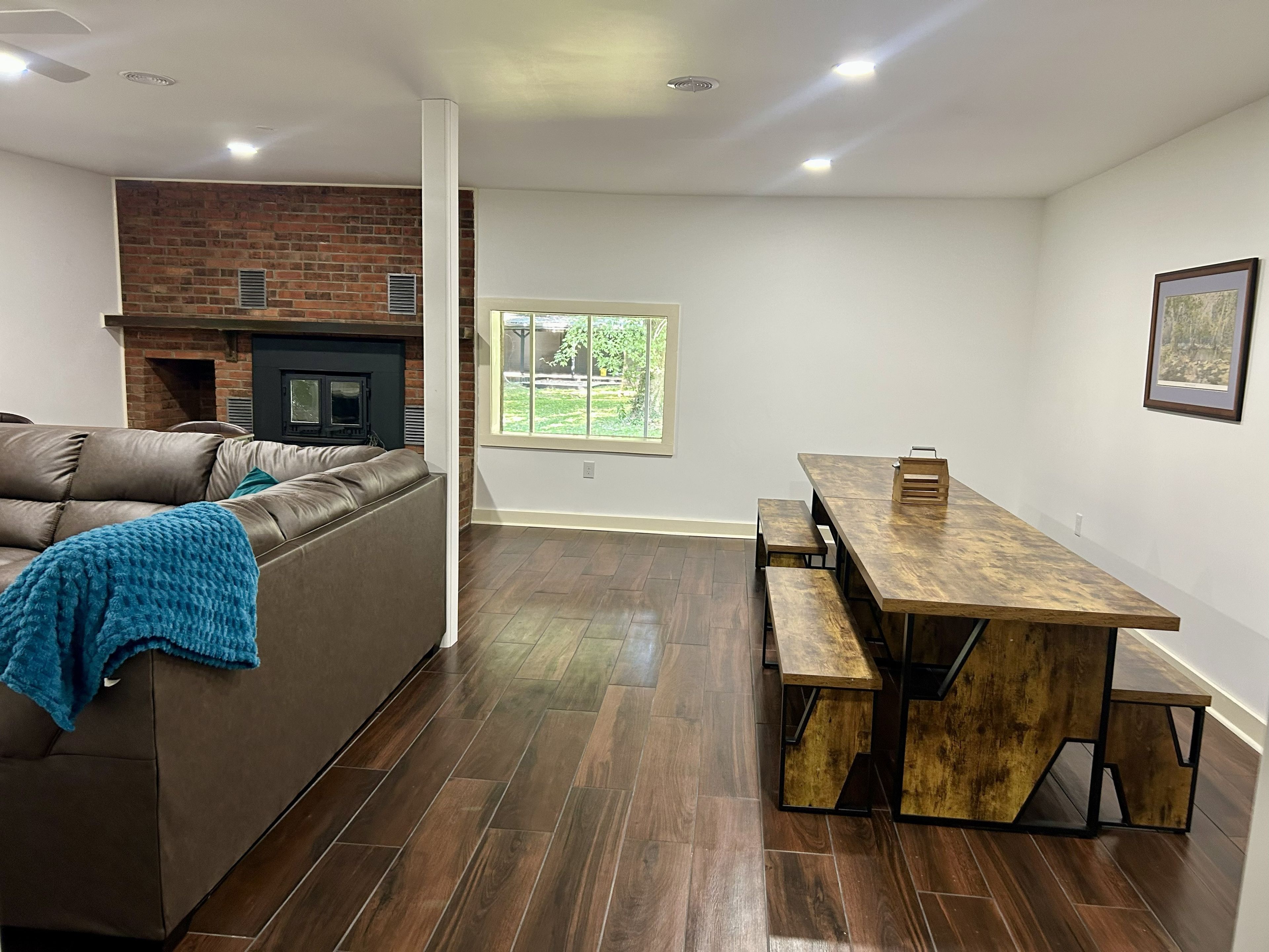 A modern living room with a leather sofa, a wooden dining table with benches, a brick fireplace, and a large window.