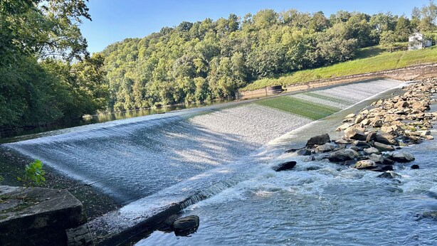 Ein Fluss fließt durch ein bewaldetes Gebiet, im Hintergrund sind ein Staudamm und ein Haus zu sehen.