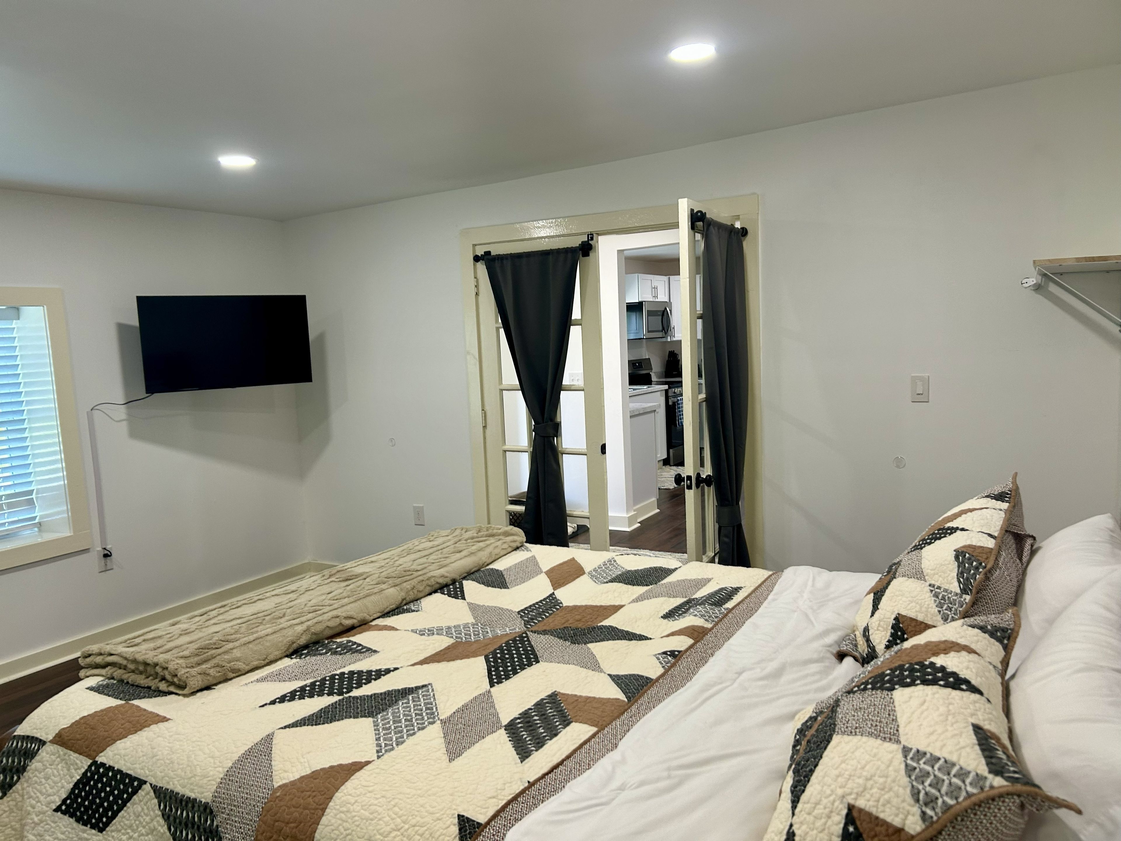 A bedroom with a bed, a TV mounted on the wall, a window with blinds, and a door leading to a kitchen.