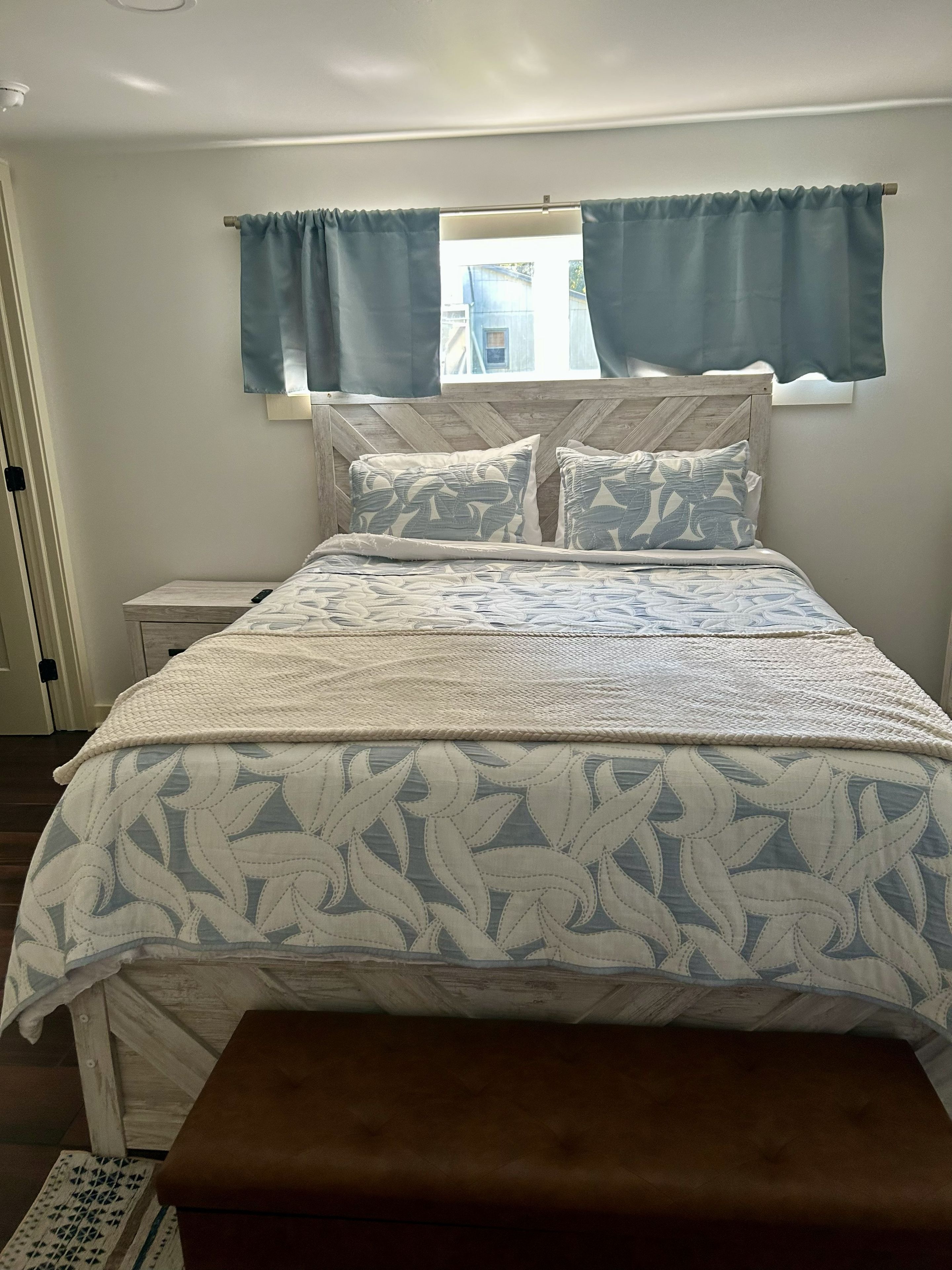 A bedroom with a bed, a bedside table, a window with curtains, and a view of the outside.