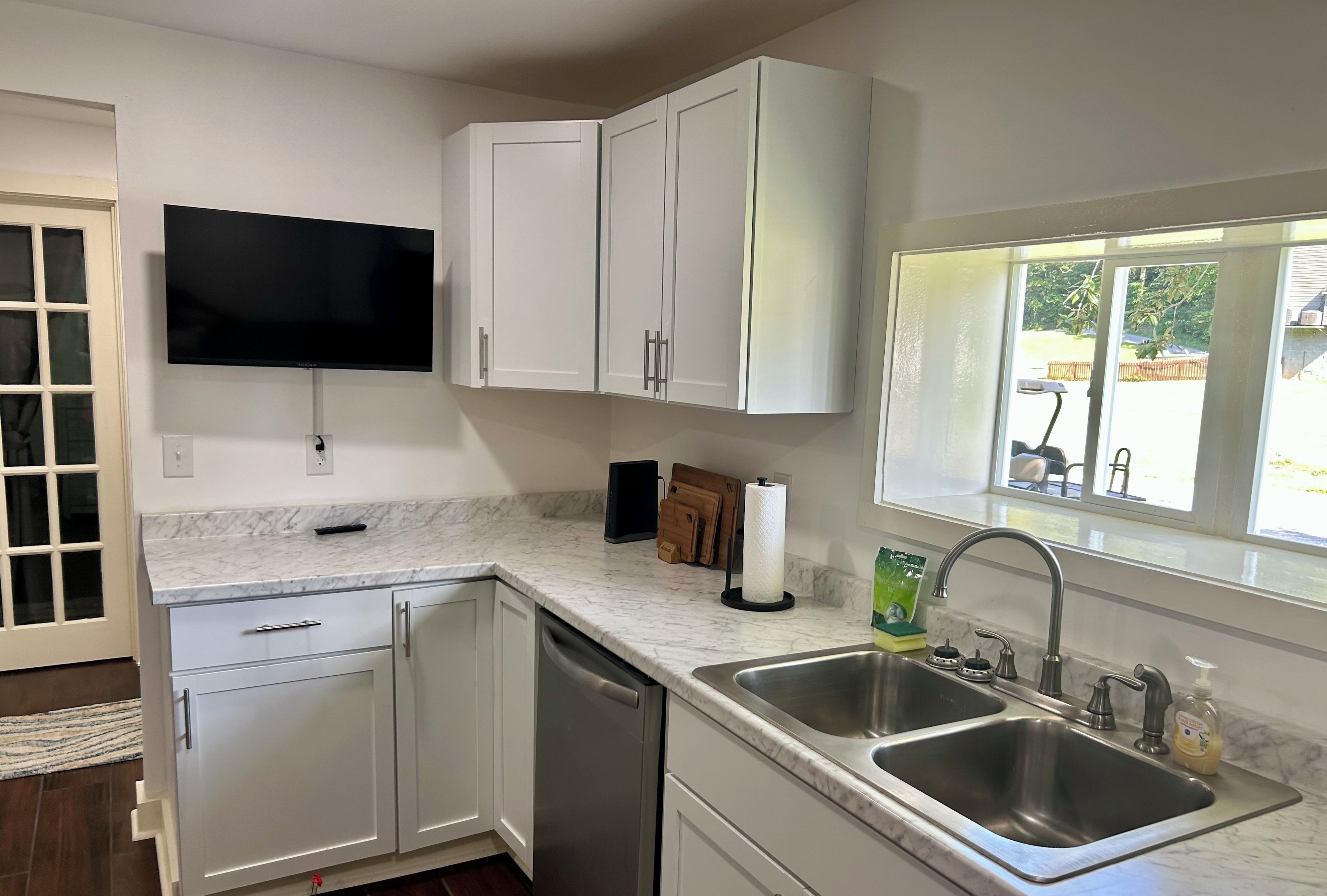 A modern kitchen with white cabinets, a built-in dishwasher, a double sink, and a flat-screen TV mounted above the countertop.