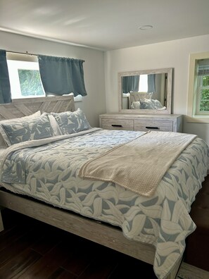 Une chambre à coucher avec un lit, une table de chevet, une commode, un miroir et une fenêtre avec des rideaux.