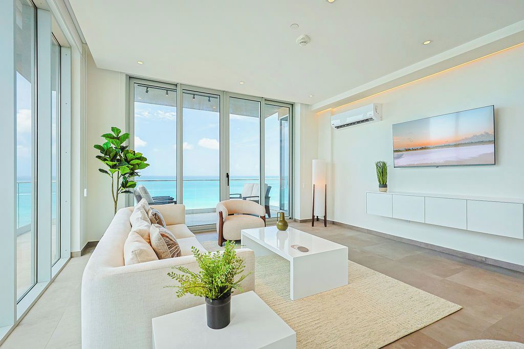 Een moderne woonkamer met een groot raam met uitzicht op zee, een flatscreen televisie aan de muur en een minimalistische meubilairindeling.