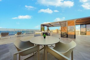 Eine Dachterrasse mit rundem Tisch und Stühlen, einem kleinen Topfpflanzen und Blick aufs Meer.