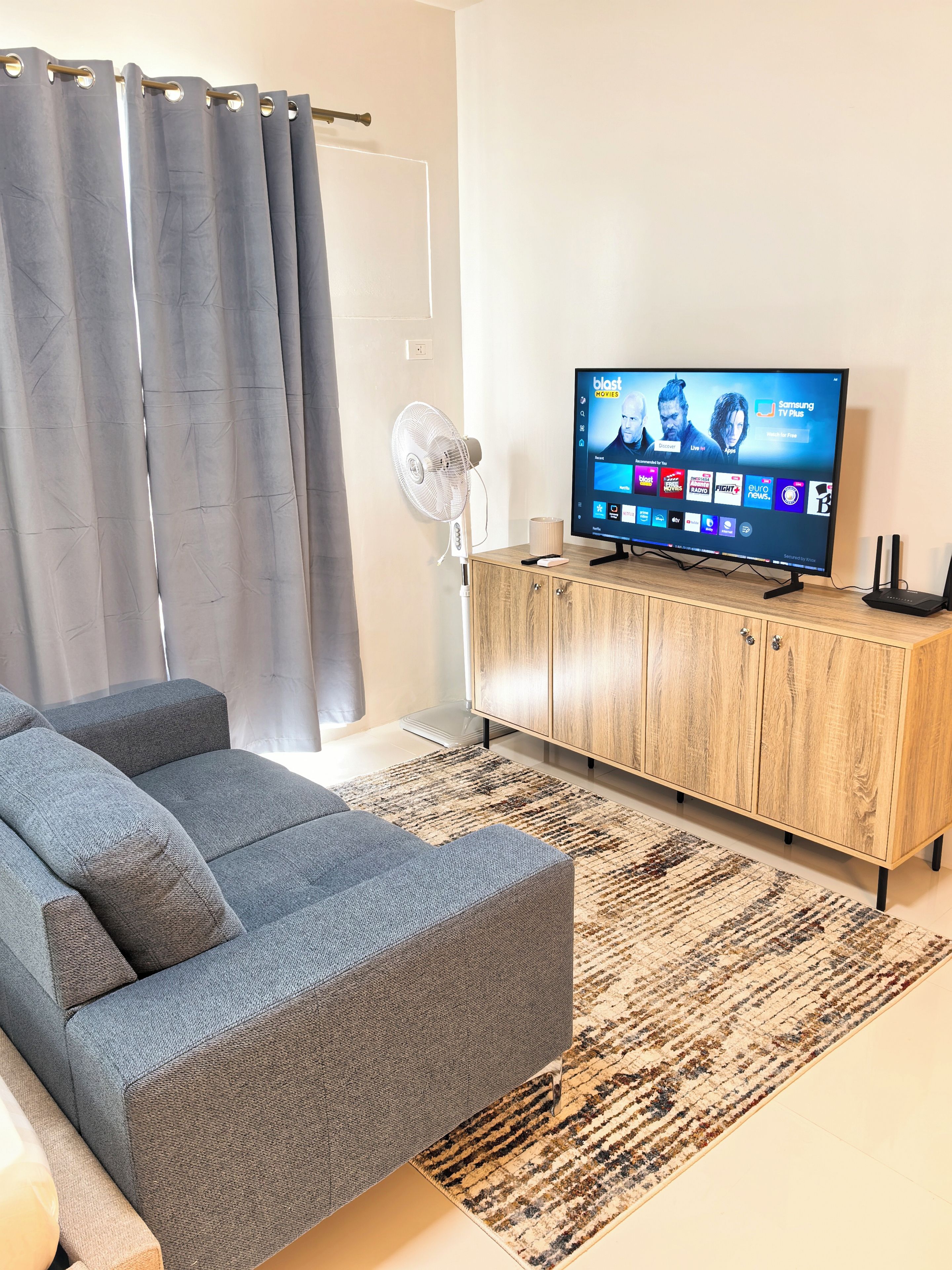 A living room with a grey sofa, a wooden cabinet, a flat-screen TV displaying a streaming service interface, and a standing fan.