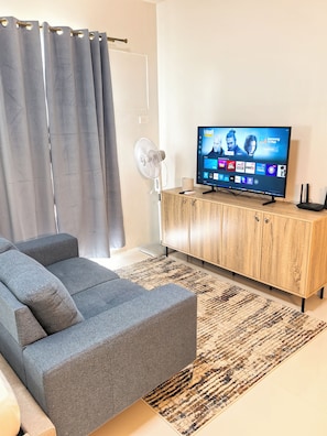 A living room with a grey sofa, a wooden cabinet, a flat-screen TV displaying a streaming service interface, and a standing fan. - Island Bliss Studio, Balcony, Pool, Gym and Near Airport (PUERTO PRINSESA)