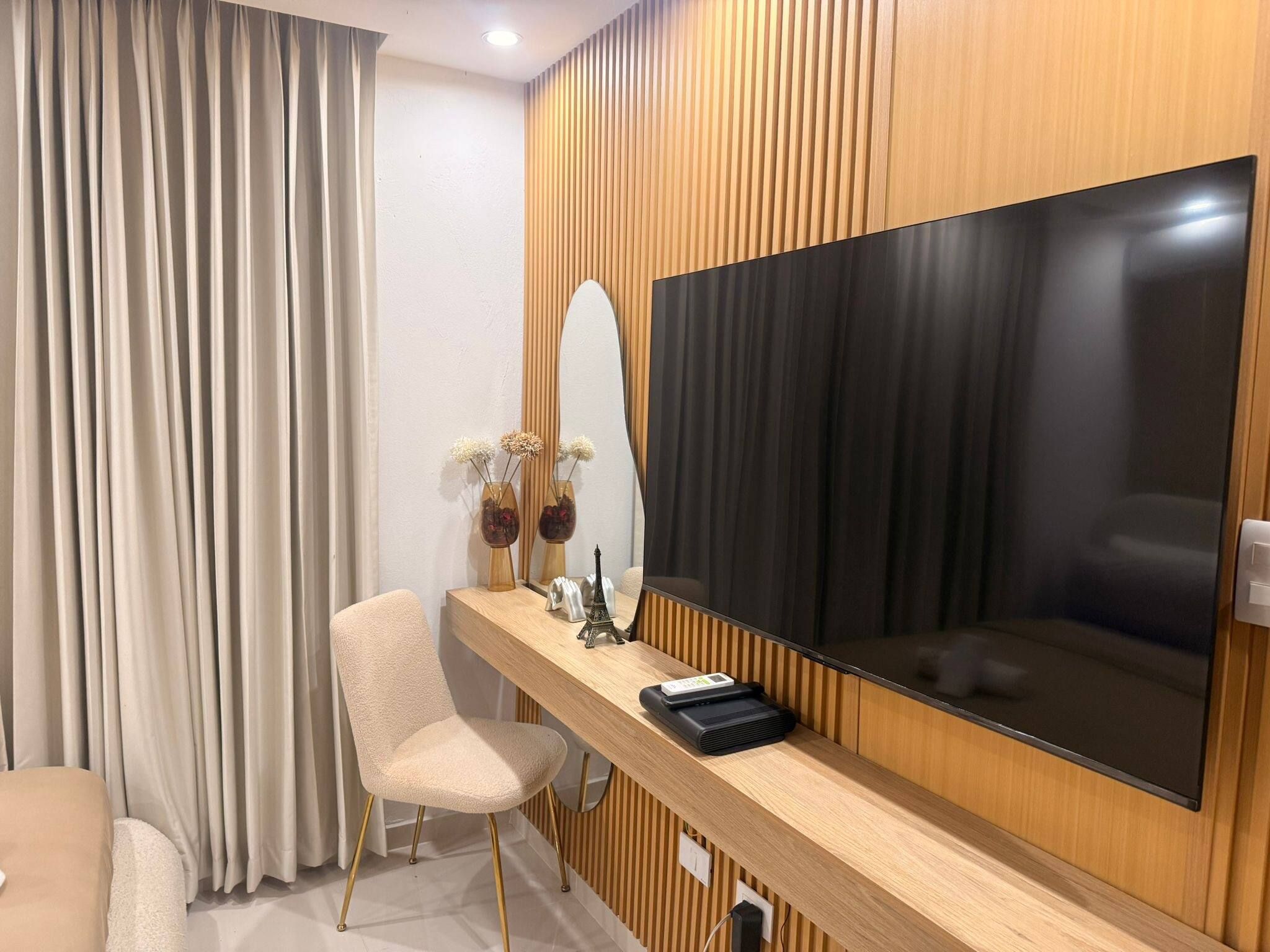 Una habitación de hotel moderna con un televisor de pantalla plana grande montado en una pared de paneles de madera, una silla cómoda, una mesita con objetos decorativos y un ventanal con cortinas.