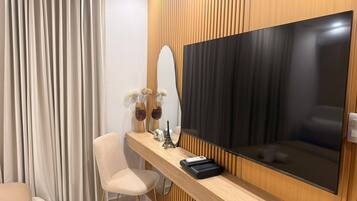 Una habitación de hotel moderna con un televisor de pantalla plana grande montado en una pared de paneles de madera, una silla cómoda, una mesita con objetos decorativos y un ventanal con cortinas.
