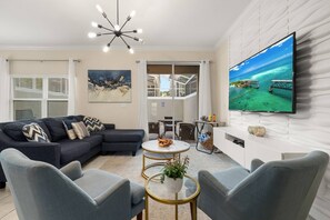 A modern living room with a sectional sofa, two armchairs, a round coffee table, a flat-screen TV mounted on the wall, and a large window with blinds. - ChampionsGate 4BR | Private Pool + Waterpark (Kissimmee)