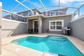 A residential pool area with a clear blue pool, a covered patio with a table and chairs, and a barbecue grill. - ChampionsGate 4BR | Private Pool + Waterpark (Kissimmee)