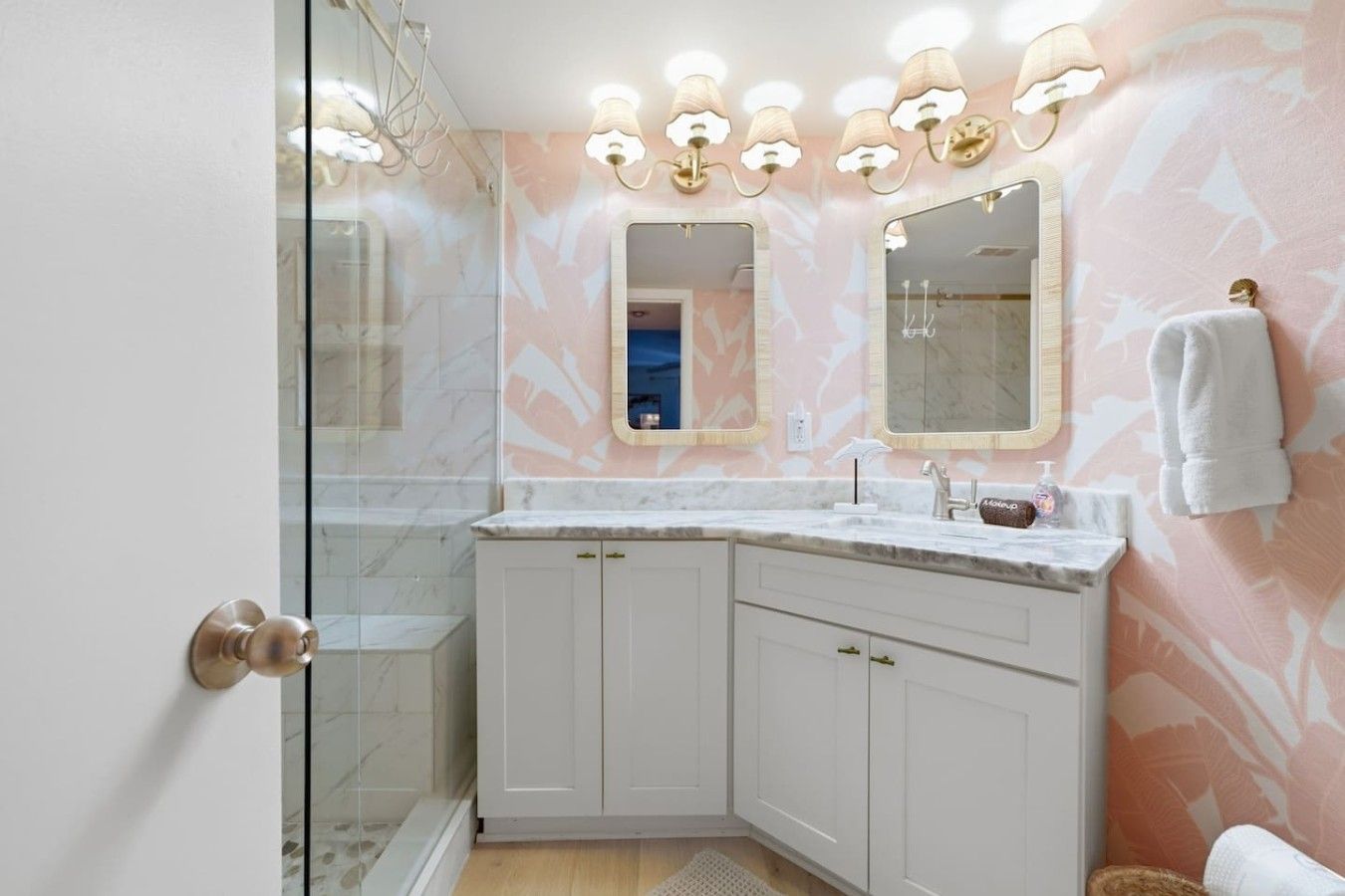 Casa de banho com cabine de duche em vidro, toucador branco com bancada de mármore e luminárias fixadas na parede.