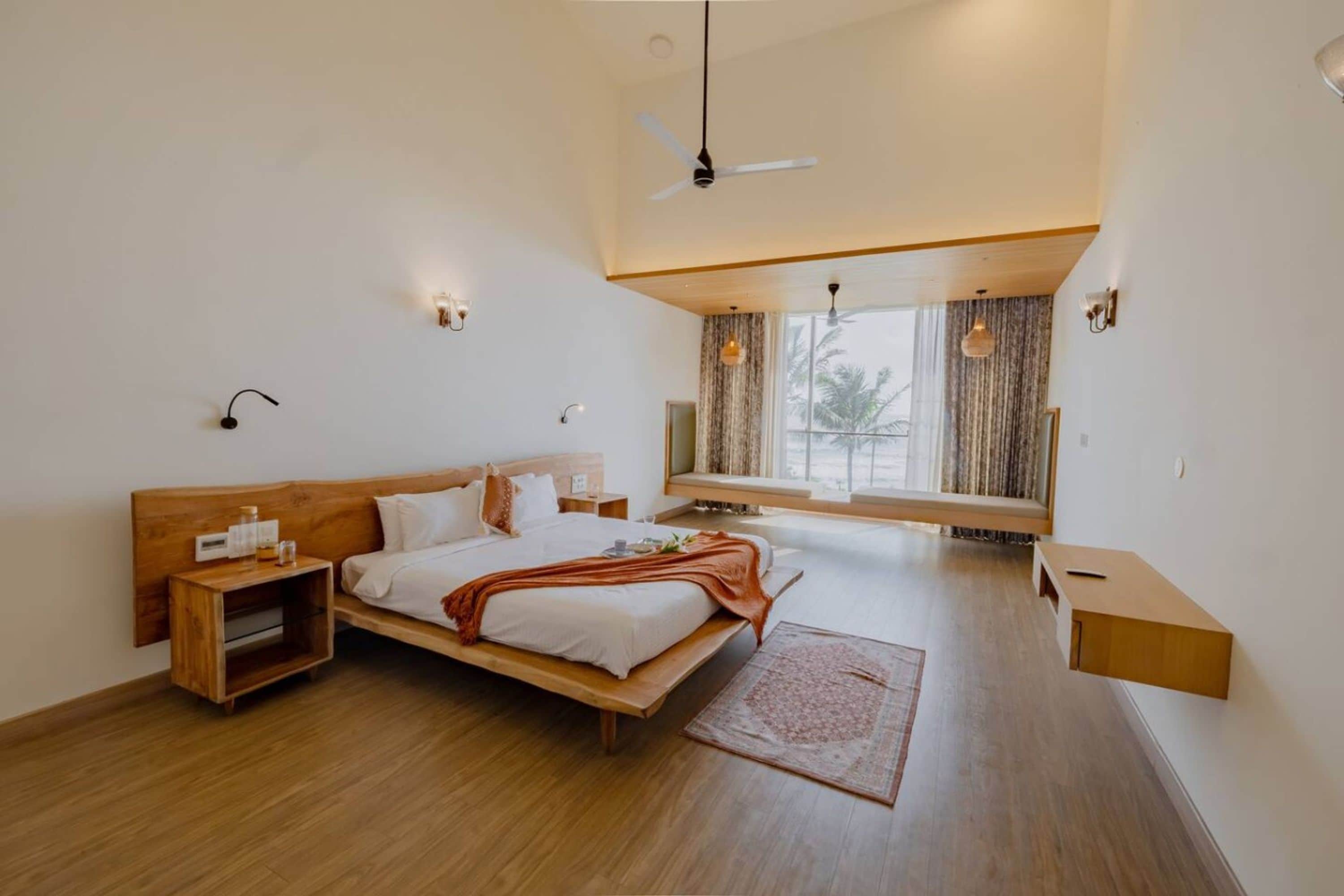 A modern bedroom with a wooden bed, bedside tables, a bench, and a view of palm trees through the window.