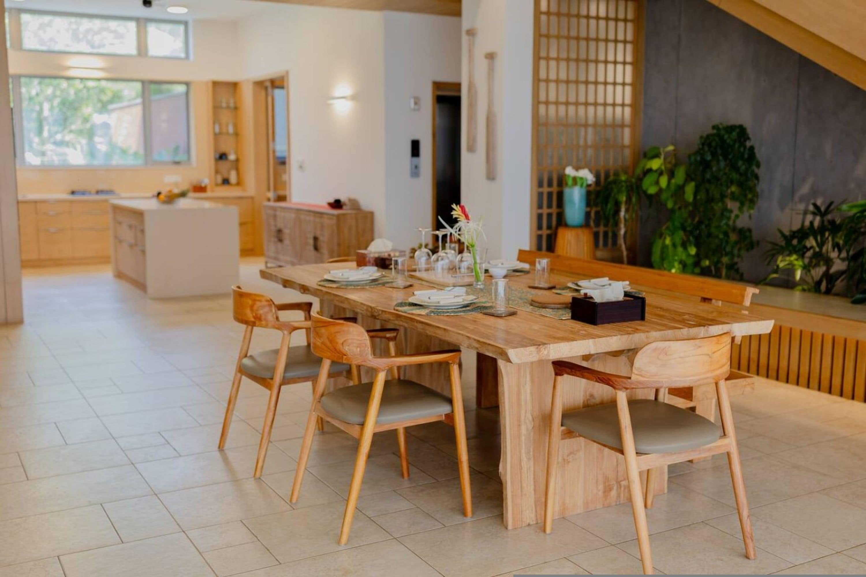 Un comedor moderno con una mesa y sillas de madera, una cocina al fondo y plantas en macetas.