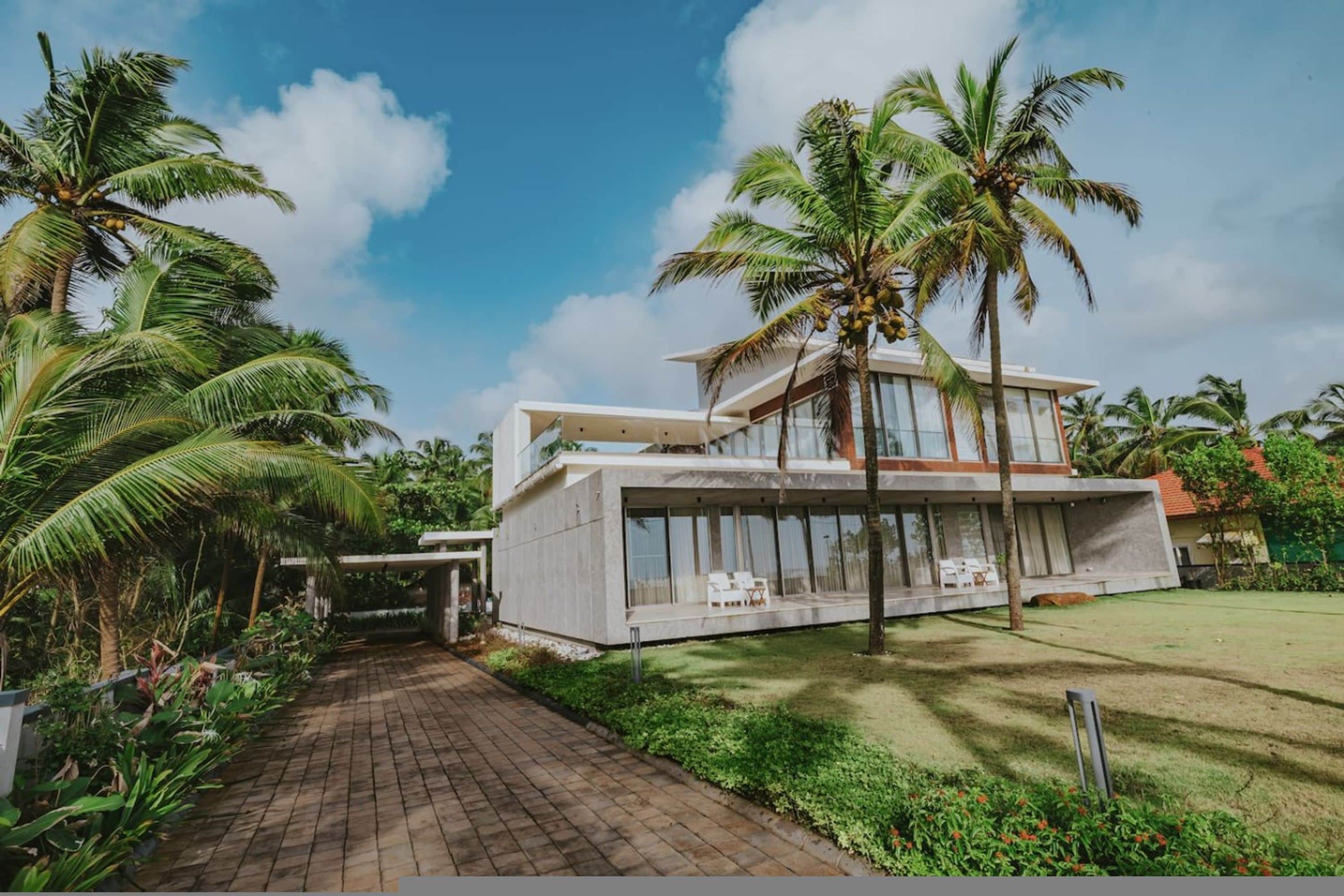 A modern two-story house with a paved pathway leading to the entrance, surrounded by lush greenery and palm trees.