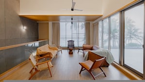A spacious room with a high ceiling, a ceiling fan, a large window with a view of palm trees, and a seating area with a sofa and chairs. - Villa Shankari - Beachside 3BR w Private Pool (Udupi)
