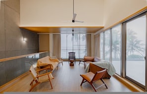 A spacious room with a high ceiling, a ceiling fan, a large window with a view of palm trees, and a seating area with a sofa and chairs. - Villa Shankari - Beachside 3BR w Private Pool (Udupi)