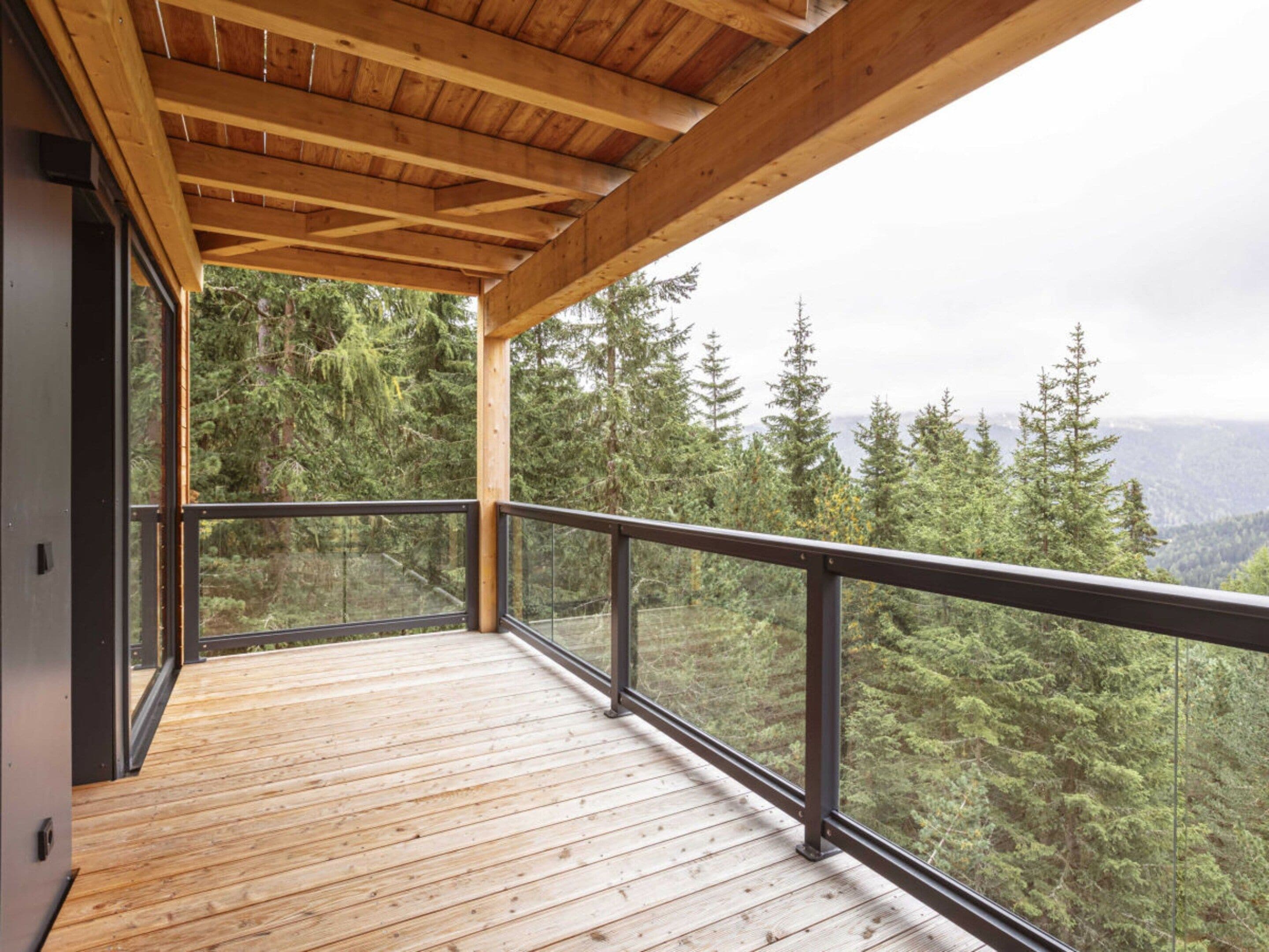 Une terrasse en bois dotée d’une balustrade en verre, offrant une vue sur un paysage boisé.