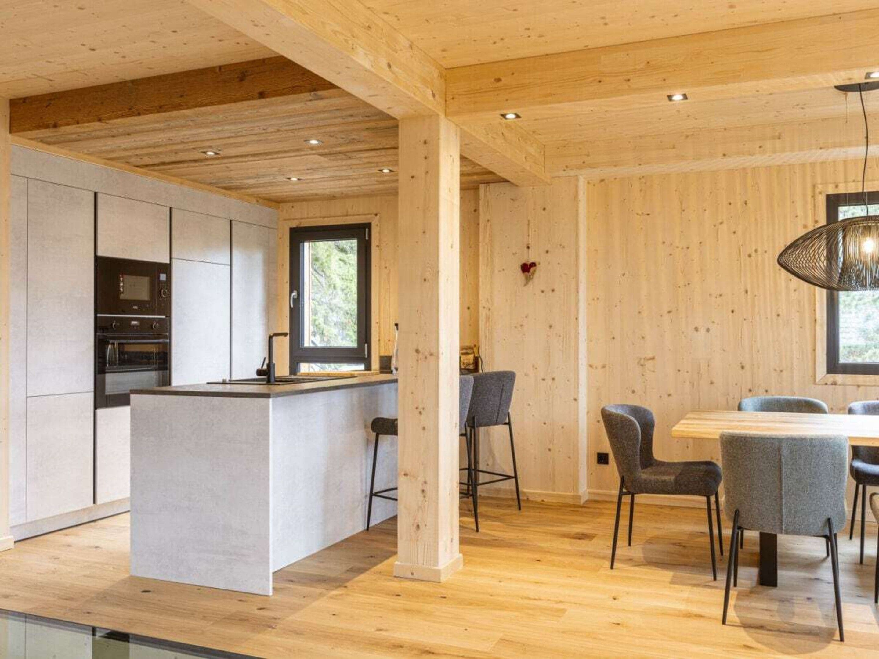 Une cuisine moderne dotée d’appareils électroménagers intégrés et d’un îlot central, un coin repas avec une table et des chaises en bois, et une grande fenêtre qui laisse entrer la lumière naturelle.