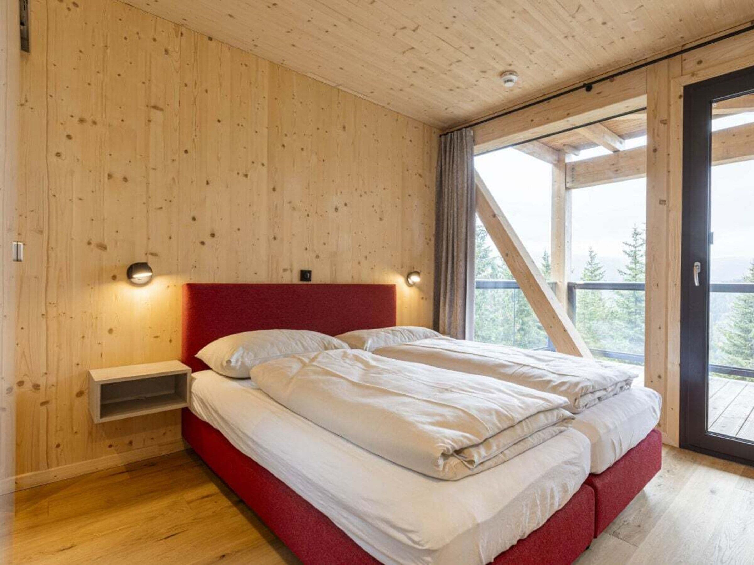 Une chambre avec un plafond en bois, une tête de lit rouge, un lit avec une literie blanche, une table de chevet et une fenêtre donnant sur des arbres.