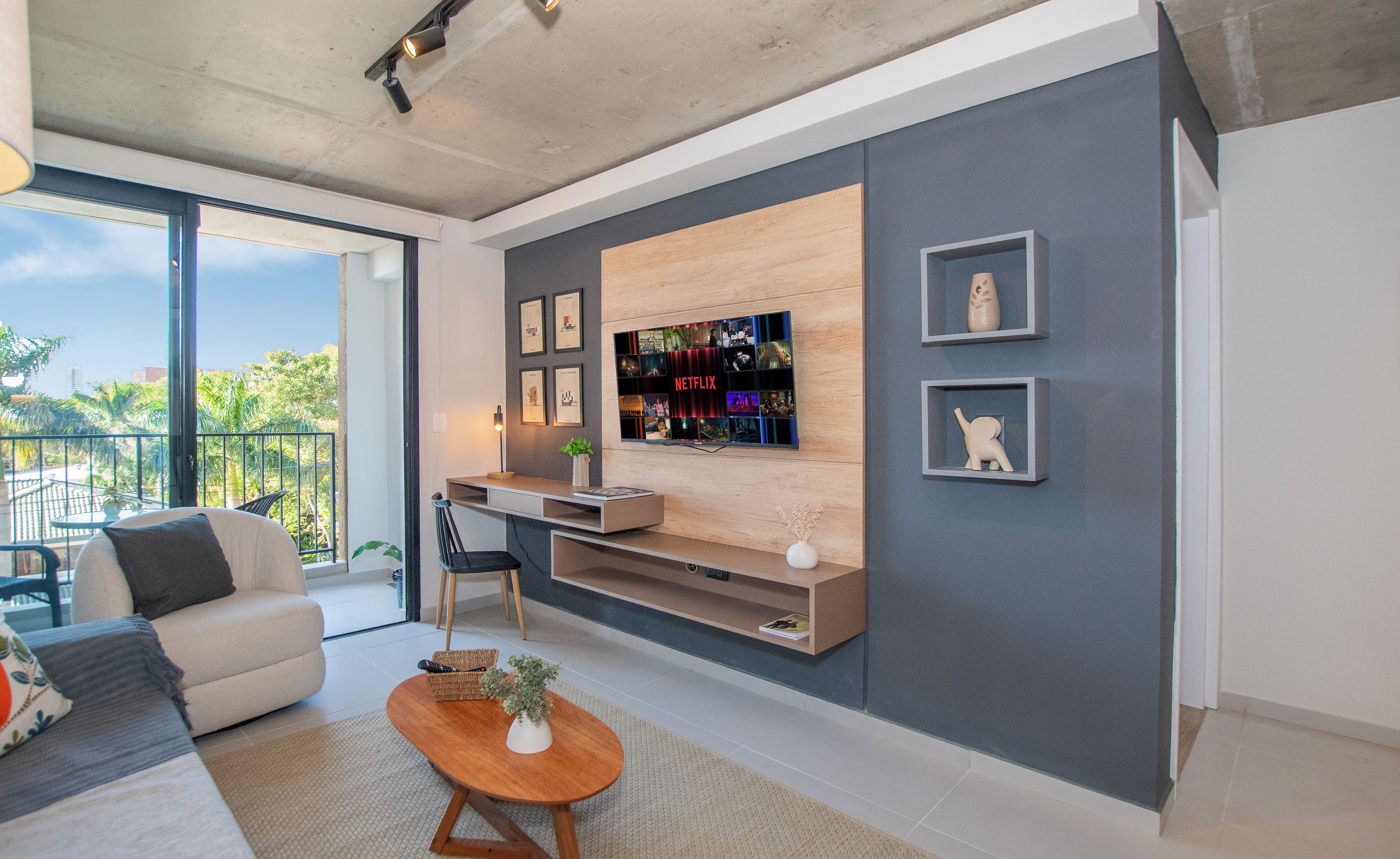 Uma sala de estar moderna com uma grande televisão de tela plana na parede, um sofá, uma mesa de centro e uma varanda com vista para palmeiras.