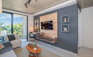 A modern living room with a large flat-screen TV mounted on a wall, a sofa, a coffee table, and a balcony with a view of palm trees. - Corporate Stays First del Sol - 301 (Asunción)
