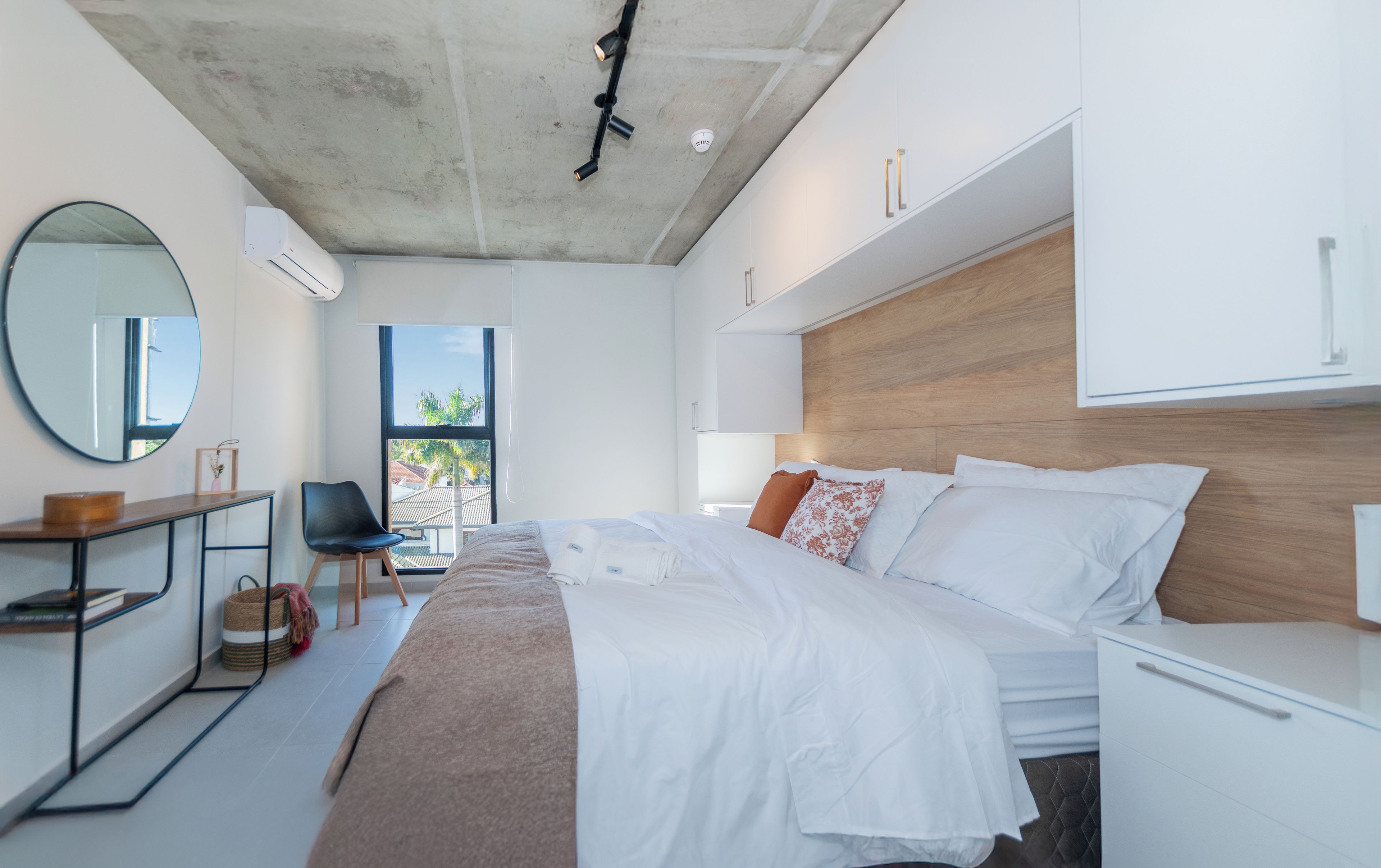 A modern bedroom with a large bed, a bedside table, a mirror, and a view of the outdoors.