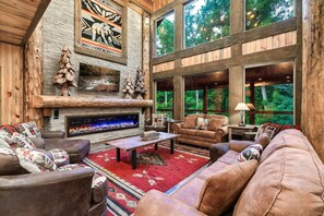 A cozy living room with a fireplace, large windows, and a view of the forest. - Luxury4bdrmCabin-LgPool-HotTub-5MinParkway-Special (Sevierville)