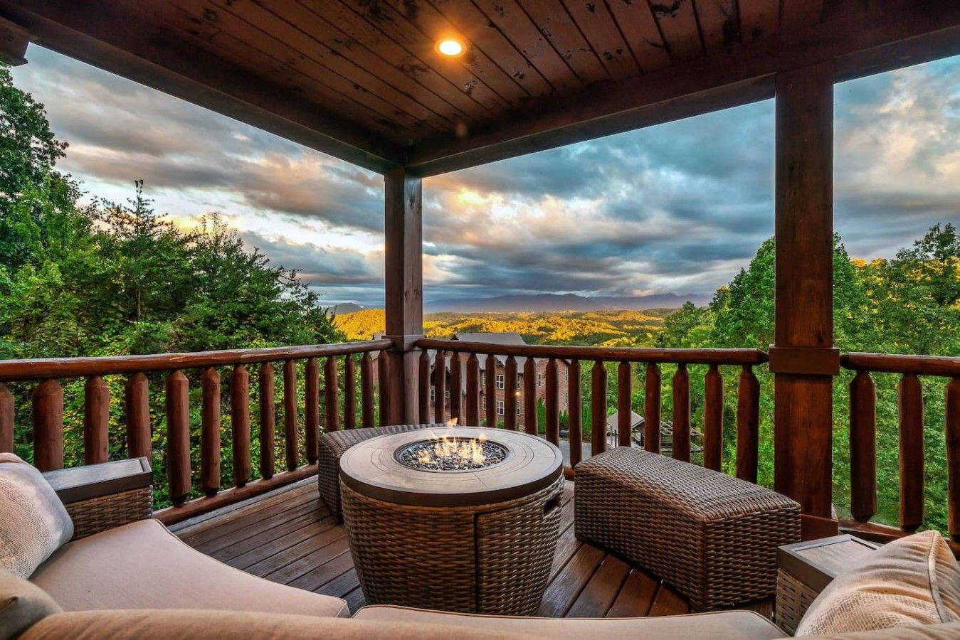 A covered outdoor deck with wicker furniture and a fire pit, overlooking a forested landscape.