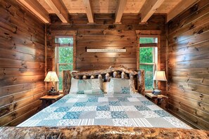 A log cabin bedroom with a rustic bed, two bedside lamps, and a view of greenery through the windows. - Indoor Golf Cabin Amazing Views Arcade 8BRDM Cabin (Sevierville)
