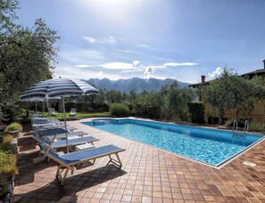 Ein Swimmingpool mit Liegestühlen und Sonnenschirmen, umgeben von viel Grün und Bergen im Hintergrund.