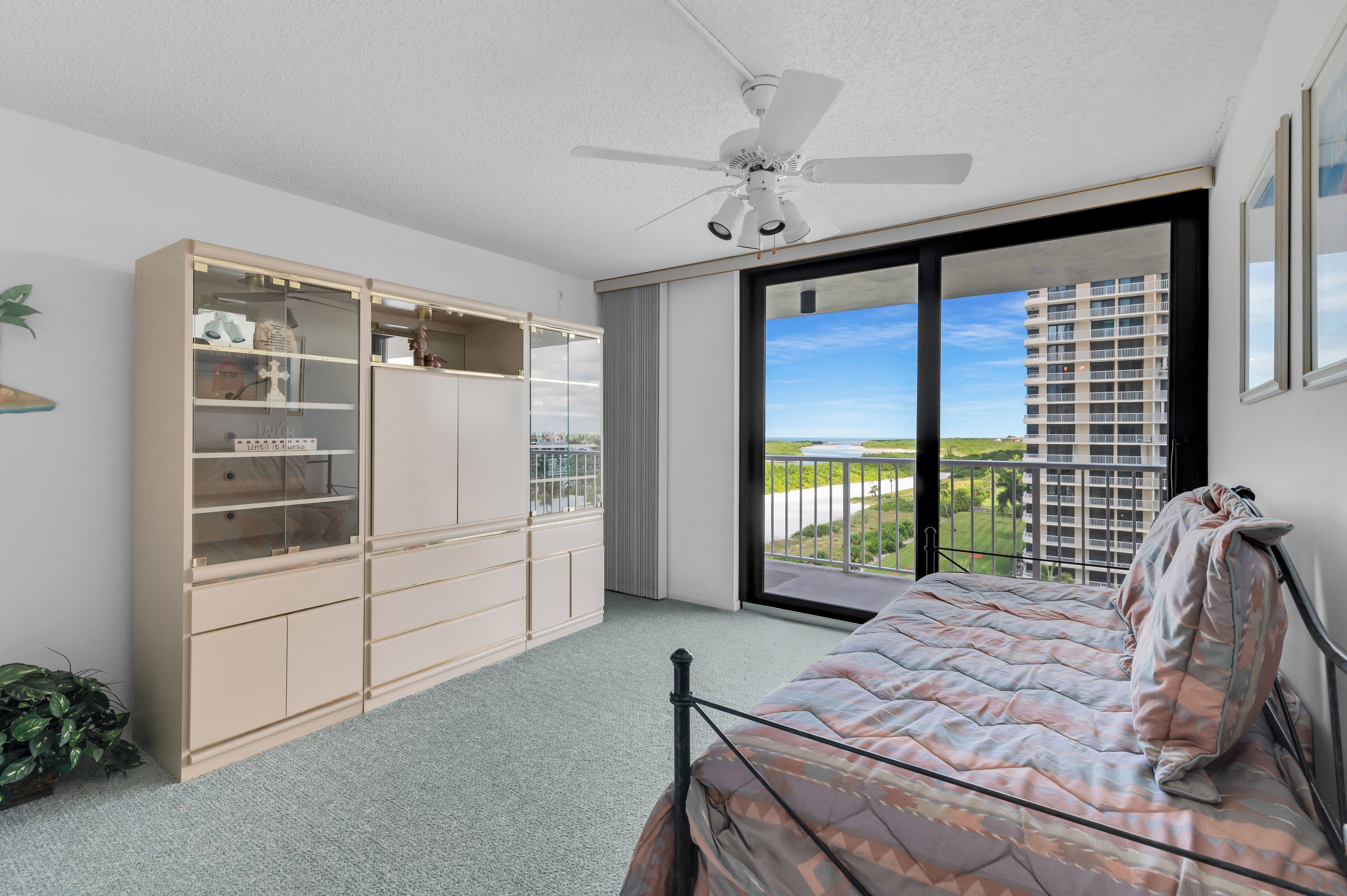 A bedroom with a bed, a wardrobe, a ceiling fan, and a large window with a view of a building and greenery.
