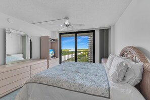 A bedroom with a bed, a ceiling fan, a television, and a balcony with a view of a building and greenery. - Beachfront | End Unit | Extended Balcony (Marco Island)