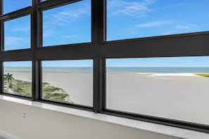 A view through a window with a clear sky and a beach in the distance. - Beachfront | End Unit | Extended Balcony (Marco Island)