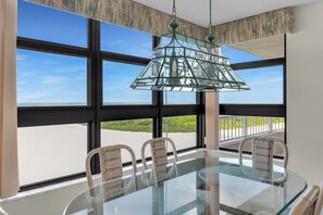 A dining area with a glass table and chairs, large windows with a view of greenery, and a decorative hanging light fixture. - Beachfront | End Unit | Extended Balcony (Marco Island)