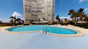 A hotel with a large swimming pool, palm trees, and a high-rise building in the background. - Beachfront | End Unit | Extended Balcony (Marco Island)