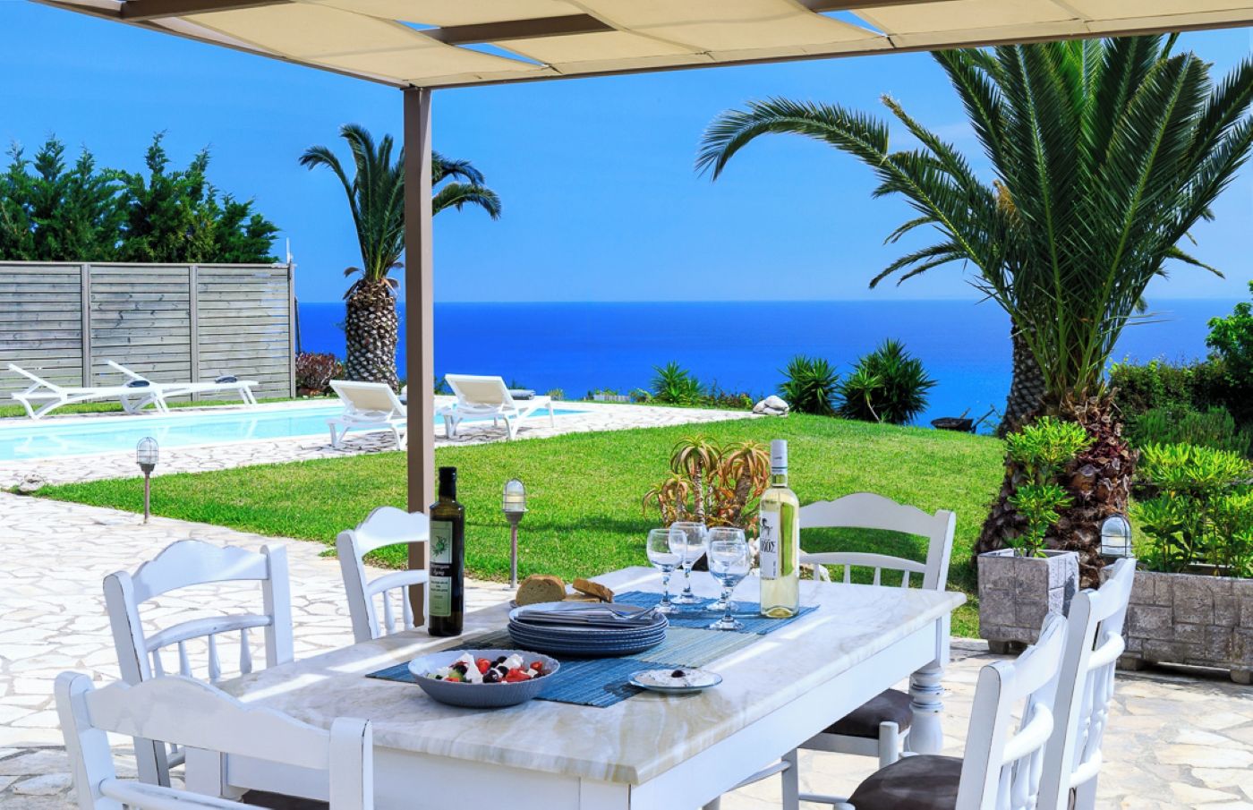 An outdoor dining area with a view of the sea, white chairs, a table set with plates, glasses, and a bottle.