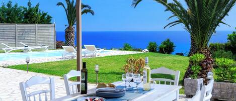 An outdoor dining area with a view of the sea, white chairs, a table set with plates, glasses, and a bottle.