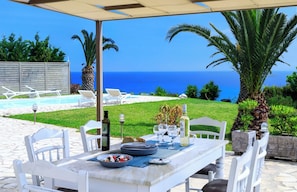 An outdoor dining area with a view of the sea, white chairs, a table set with plates, glasses, and a bottle. - Villa Mariza, a laid-back hillside villa with panoramic views and beautiful gardens (Kefalonia)