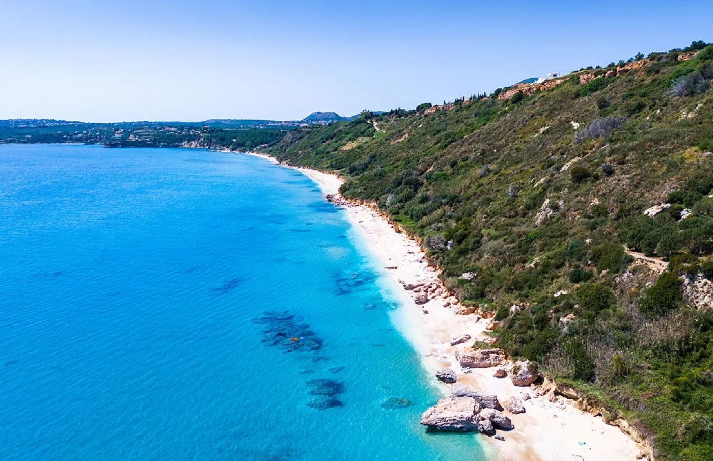 Paisagem costeira com mar azul e límpido, praia arenosa e relevo montanhoso com vegetação escassa.
